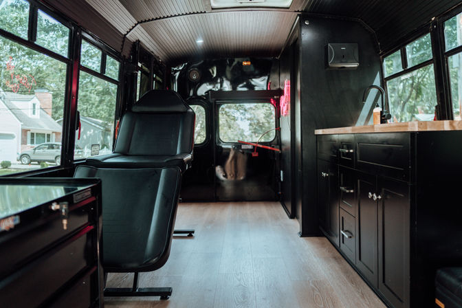 Cozy converted bus mobile studio interior with a black reclining chair, wood floors, matte black cabinets and sink, and large windows overlooking a suburban street.