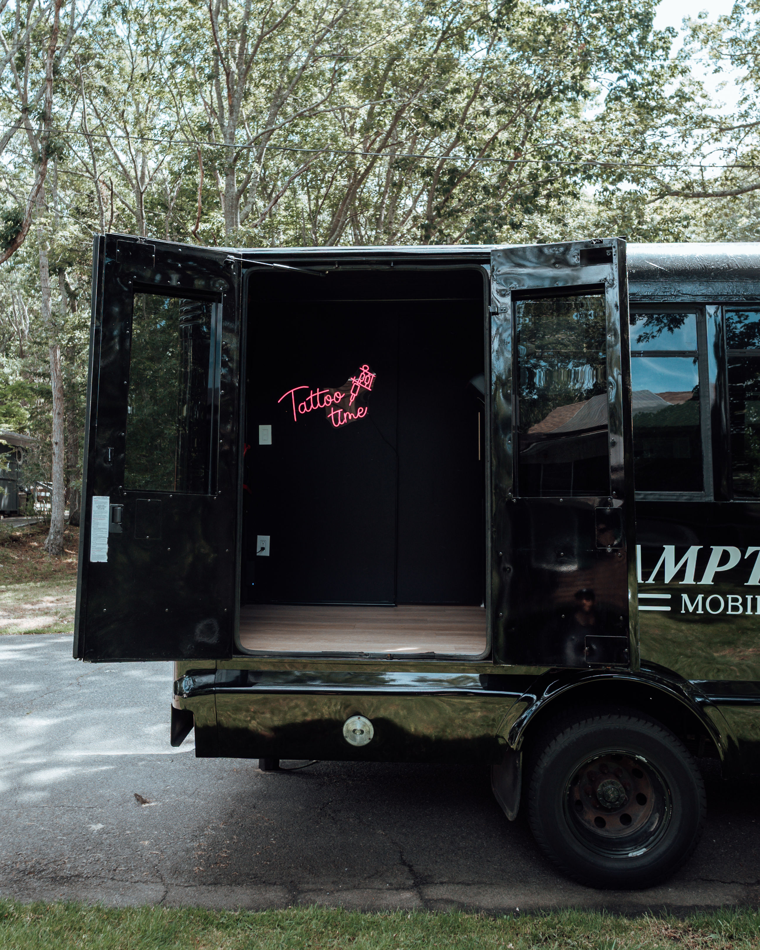 Black mobile tattoo van parked on a tree-lined suburban street with rear doors open, revealing a pink neon “Tattoo time” sign inside — mobile tattoo studio setup.