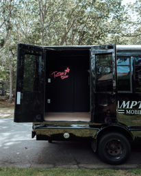 Black mobile tattoo van parked on a tree-lined suburban street with rear doors open, revealing a pink neon “Tattoo time” sign inside — mobile tattoo studio setup.