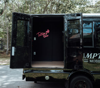 Black mobile tattoo van parked on a tree-lined suburban street with rear doors open, revealing a pink neon “Tattoo time” sign inside — mobile tattoo studio setup.