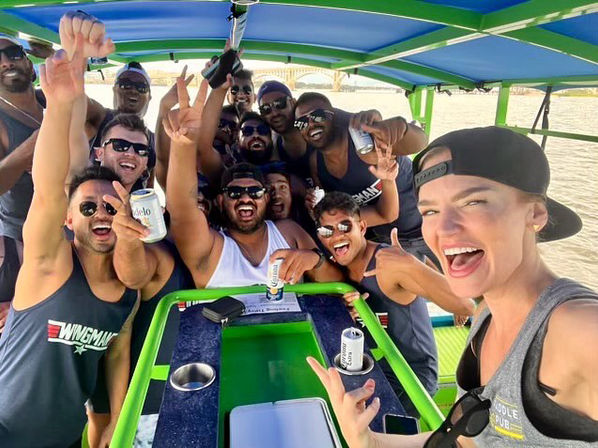Group selfie of people cheering with canned drinks on a green-and-blue party boat by a sandy beach and shoreline bridge on a sunny day