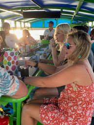Group of friends enjoying drinks at a colorful floating bar on a boat at sunset, green counter and blue canopy, cans and tumblers on the bar, lively summer waterfront vibe.