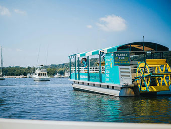 Turquoise pedal-powered paddlewheel boat with a bright yellow wheel cruising a calm harbor near anchored sailboats and a tree-lined shoreline under a clear blue summer sky.