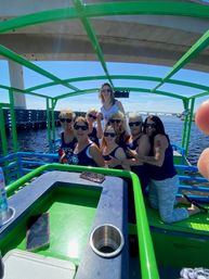 Group of women in sunglasses posing and smiling on a bright green party boat under a concrete bridge on a sunny river, with a distant arched bridge and waterfront in the background.
