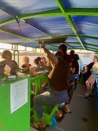 Group of people pedaling and chatting on a bright green pedal-bar boat under a blue canopy at sunset, waterfront and bridge visible in the background.