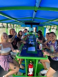 Group of smiling adults enjoying drinks on a bright blue-and-green open-air party trolley during a daytime outdoor ride.