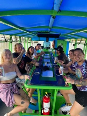Group of smiling adults enjoying drinks on a bright blue-and-green open-air party trolley during a daytime outdoor ride.