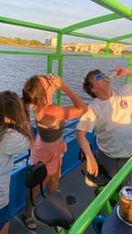 People on a green-framed river party boat at sunset, standing by bike-style seats and chugging canned drinks with water and an arched bridge in the background.
