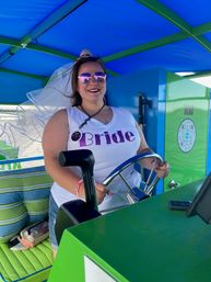 Smiling bride in glitter "Bride" tank top, veil and pink mirrored sunglasses steering a bright green-and-blue party boat — bachelorette on the water