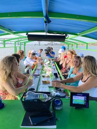 Passengers on a green-framed pontoon boat with a blue canopy, enjoying drinks and snacks at a long center table while a cameraman films; calm river and arched bridge in the sunny background.