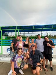 Cheerful group of friends posing on a colorful green-and-blue tour boat dock, wearing casual summer clothes and smiling beneath a faint rainbow in a cloudy sky.