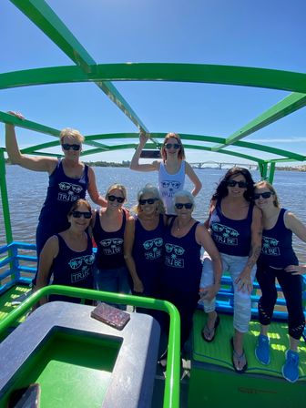 Group of women wearing matching navy TRIBE tank tops and sunglasses posing on a bright green-framed open-top boat during a sunny river cruise, calm water and an arched bridge visible on the horizon.