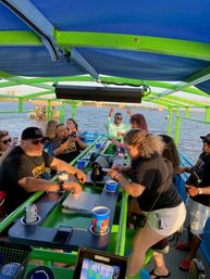 Friends enjoying drinks at a bright green covered party-boat bar during a sunset river cruise, with a bridge and waterfront in the background.