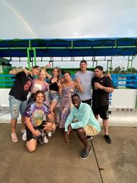 Cheerful group of eight friends posing on a waterfront pier under a blue-green canopy, casual summer outfits and a faint rainbow arching across a cloudy sky.