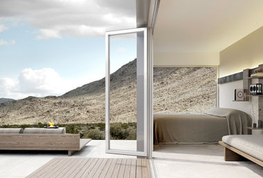 Minimalist modern desert villa with open glass door from bedroom to wooden patio, outdoor fire pit and rocky desert hills under a bright blue sky