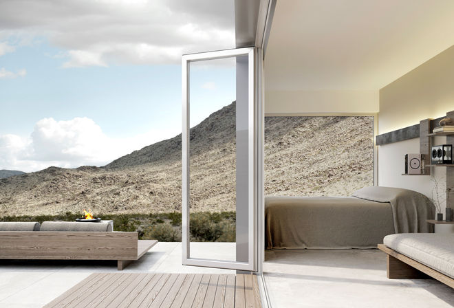 Minimalist modern desert villa with open glass door from bedroom to wooden patio, outdoor fire pit and rocky desert hills under a bright blue sky