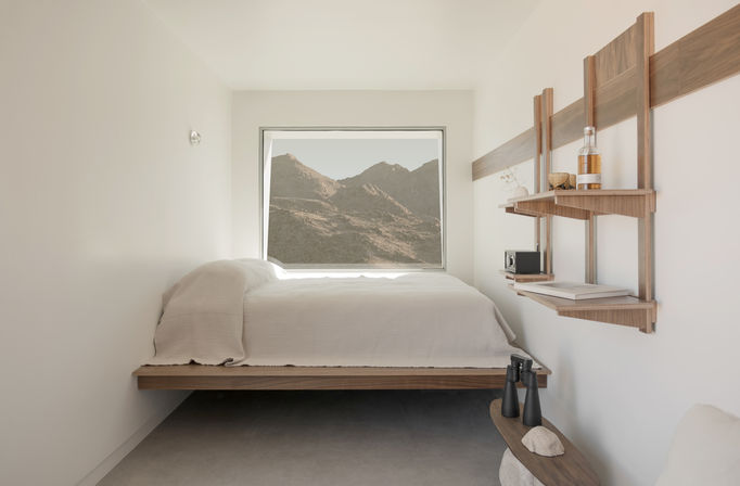 Sunlit minimalist mountain-view bedroom with a floating wood platform bed dressed in white linens, a large window framing rugged peaks, wooden wall shelves with books and a bottle, and a small bench with binoculars and decor.