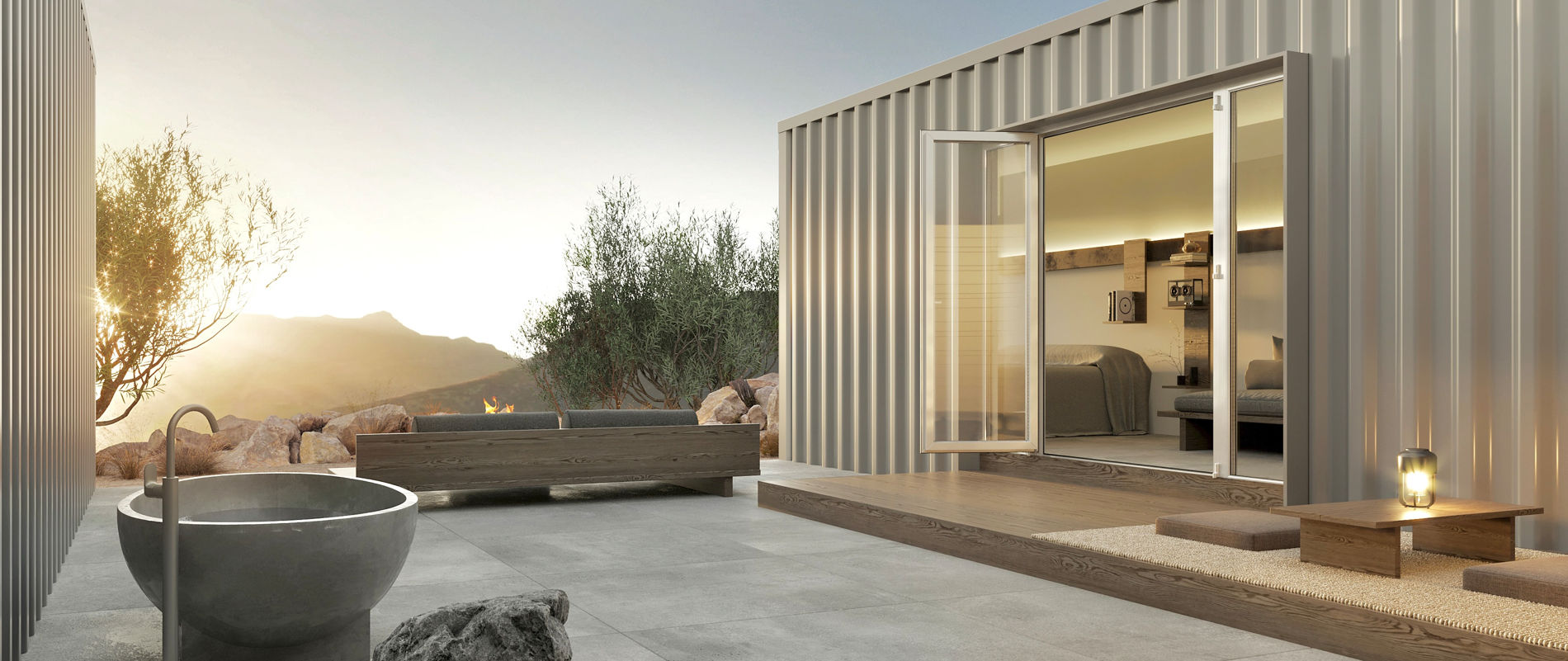 Modern container-style cabin patio at sunset with open glass doors to a cozy bedroom, wooden deck seating and low table with lantern, concrete outdoor soaking tub, fire pit and rugged mountain view.