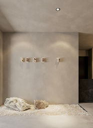 Modern minimalist lobby interior with beige plaster wall featuring raised 'RESET' letters, two large rocks on white gravel and warm recessed ceiling light — zen design.