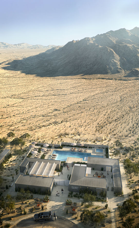 Aerial view of a modern desert oasis with modular low buildings and a rectangular outdoor pool surrounded by lounge chairs, set in a sandy arid valley backed by rugged mountains under a clear blue sky.