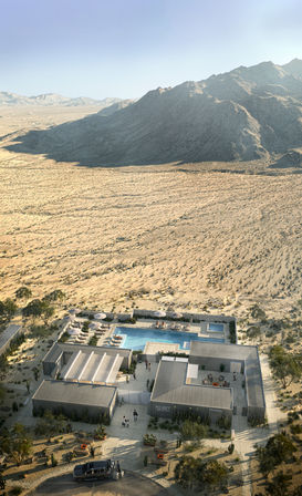 Aerial view of a modern desert oasis with modular low buildings and a rectangular outdoor pool surrounded by lounge chairs, set in a sandy arid valley backed by rugged mountains under a clear blue sky.