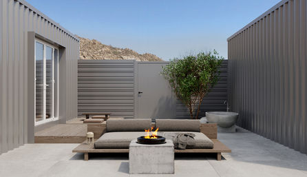 Minimalist container-style courtyard patio with a low wooden lounge sofa, lit round fire pit on a concrete block, potted tree and stone basin, with rocky hills under a clear blue sky.