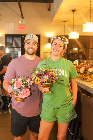 Smiling man and woman holding gold compote vases filled with pink and yellow mixed-flower arrangements in a warm-lit floral workshop or café with hanging pendant lights.