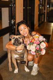 Smiling woman crouching in an indoor evening setting, hugging a large tan dog in a harness and holding a vibrant mixed bouquet of roses and seasonal flowers.