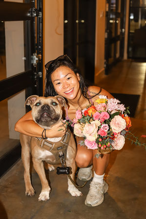 Smiling woman crouching in an indoor evening setting, hugging a large tan dog in a harness and holding a vibrant mixed bouquet of roses and seasonal flowers.