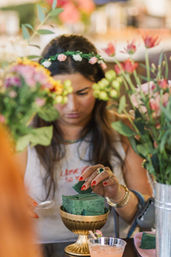 Person wearing a floral crown placing green floral foam into a gold compote surrounded by colorful blooms at an indoor DIY flower-arranging workshop
