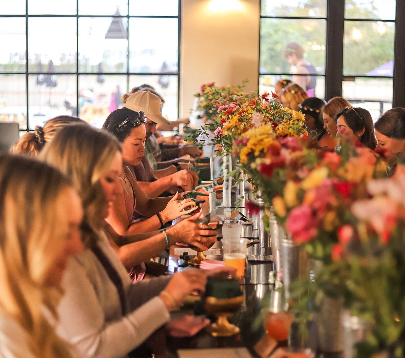 Group flower-arranging workshop at a long communal table, people crafting colorful floral bouquets and sipping drinks by large grid windows