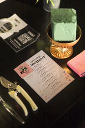 Pet-themed floral workshop station on a black table: green floral foam in a gold bowl, pruning shears, pink towel, and a DIY workshop worksheet with event cards.