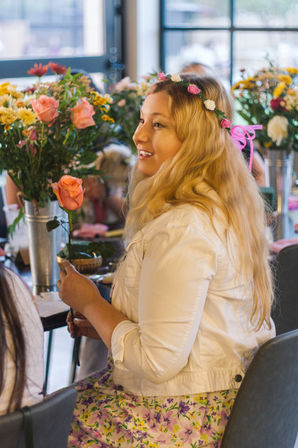 Smiling woman with long blonde hair wearing a floral crown and white jacket holds a pink rose at an indoor floral workshop surrounded by colorful bouquets.