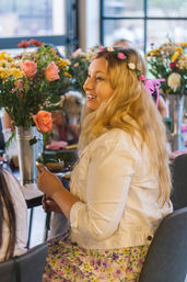 Smiling woman wearing a flower crown and white jacket holds a single pink rose while seated at a table surrounded by bouquets in an indoor floral workshop.