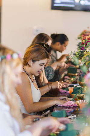 Group of women at an indoor floral workshop, hands-on arranging bouquets on a long table with colorful blooms and green floral foam.