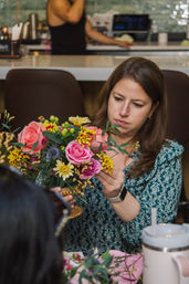Person arranging a colorful bouquet of pink roses, yellow chrysanthemums and greenery in a gold compote vase at a cozy indoor workshop table.