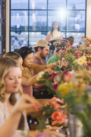 Hands-on flower-arranging workshop in a bright studio, people seated along a long communal table crafting colorful bouquets and stems.
