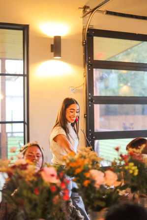 Beaming host with headset microphone leading a flower-arranging workshop in a modern indoor event space, colorful blooms in the foreground and warm lighting