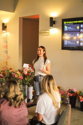 Smiling woman presenter wearing a headset leads a small indoor workshop, colorful flower arrangements on the table, seated attendees listening, wall lights and a TV leaderboard display visible in the background.