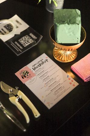 Indoor floral workshop table: green floral foam block in a gold compote, pruning shears, pink towel, and laminated instruction sheet.