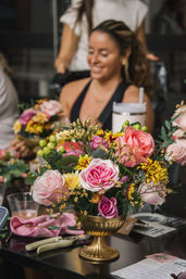 Cheerful gold urn centerpiece with pink and coral roses, yellow chrysanthemums, green berries and greenery on a table during an indoor flower-arranging workshop, blurred smiling woman in the background.