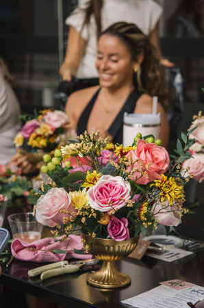 Cheerful gold urn centerpiece with pink and coral roses, yellow chrysanthemums, green berries and greenery on a table during an indoor flower-arranging workshop, blurred smiling woman in the background.