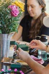 Hands placing green floral foam in a gold compote during an indoor DIY flower-arranging workshop, with a metal bucket of pink roses and yellow daisies and a blurred participant in the background.