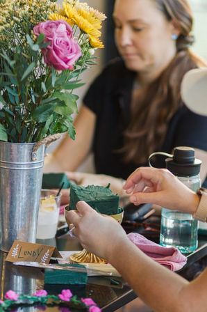 Hands placing green floral foam in a gold compote during an indoor DIY flower-arranging workshop, with a metal bucket of pink roses and yellow daisies and a blurred participant in the background.