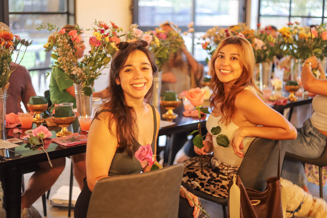 Two smiling women holding roses at an indoor flower-arranging workshop, seated at a long table filled with vases of colorful blooms and warm golden light.