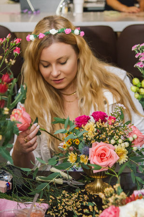 Blonde woman wearing a flower crown arranging a colorful bouquet of pink roses, hot-pink blooms, yellow chrysanthemums and eucalyptus in a gold vase at a floral workshop.