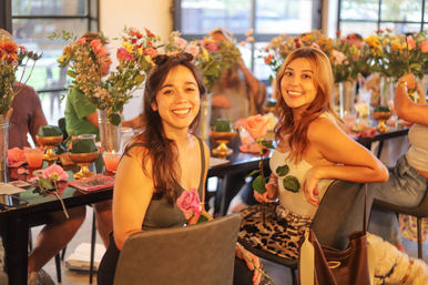 Two smiling women holding roses at an indoor flower-arranging workshop, seated at a long table decorated with vases of mixed blooms, cocktails, and gold accents
