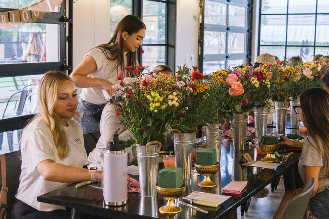 Women at an indoor floral workshop arranging colorful bouquets along a long communal table with metal buckets and gold bowls in a bright glass-walled studio.