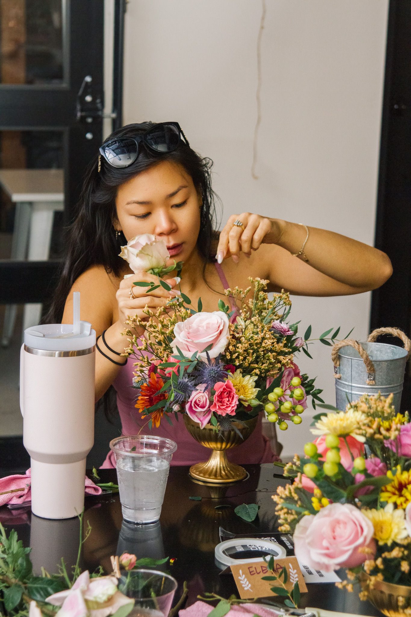 Woman arranging a pink-rose floral centerpiece in a gold compote at an indoor DIY flower workshop, table with tumbler, water cup and floristry tools.