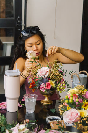 Woman arranging a pink-rose floral centerpiece in a gold compote at an indoor DIY flower workshop, table with tumbler, water cup and floristry tools.
